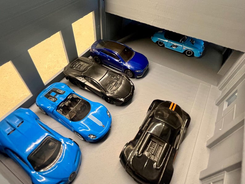 May include: A collection of toy cars in various colors, including blue and black, are arranged in a miniature garage setting. The cars have detailed features, such as headlights and racing stripes.