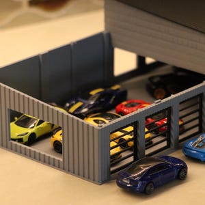 May include: A miniature gray garage with several toy cars inside and outside. The cars are various colors, including yellow, blue, red, and black. The garage has a flat roof and a few open bays.