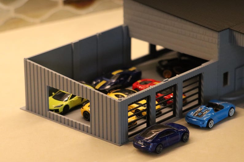 May include: A miniature gray garage with several toy cars inside and outside. The cars are various colors, including yellow, blue, red, and black. The garage has a flat roof and a few open bays.