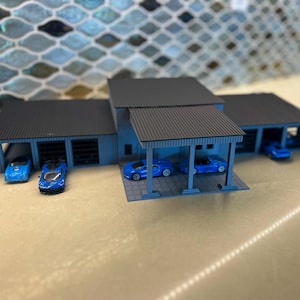 May include: Miniature blue car models parked in a light blue garage with a black roof. The garage has multiple bays and a covered carport area. The background features a mosaic tile pattern.