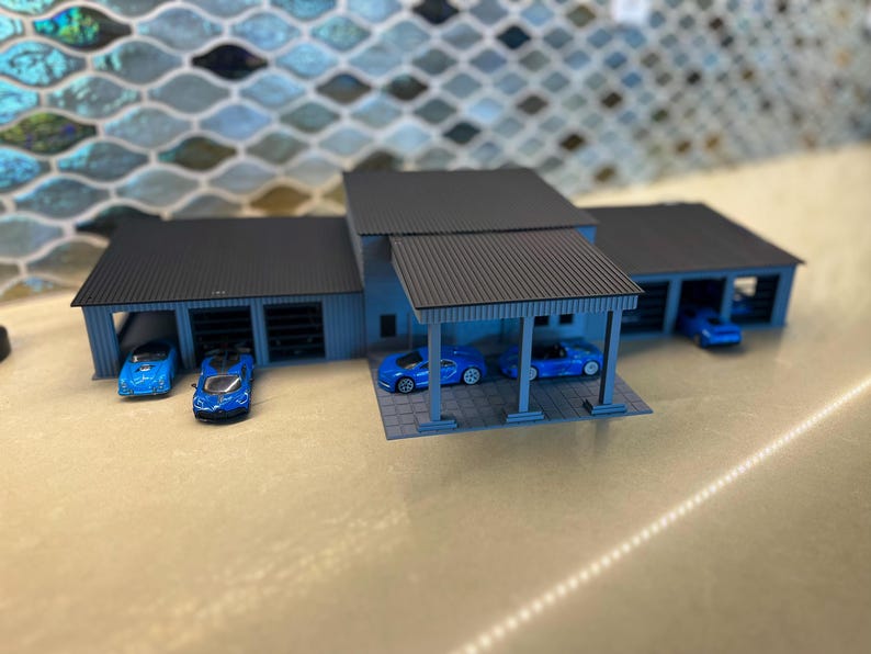 May include: Miniature blue car models parked in a light blue garage with a black roof. The garage has multiple bays and a covered carport area. The background features a mosaic tile pattern.