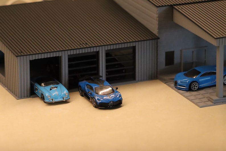 May include: Miniature blue sports cars parked in and around a model garage. The garage has three bays, with two cars inside and one parked outside. The cars are various shades of blue.