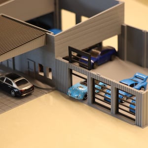 May include: A miniature model of a modern garage with several toy cars. The garage has a carport and multiple bays, each housing a different colored car, including blue and silver vehicles. The structure is gray with a brown roof.