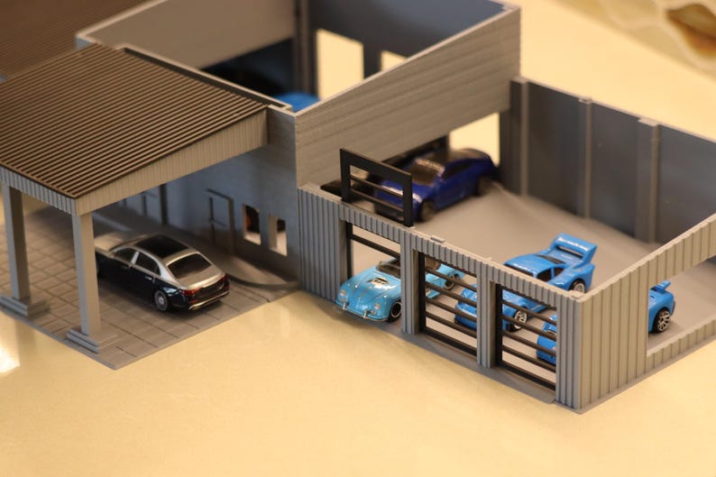 May include: A miniature model of a modern garage with several toy cars. The garage has a carport and multiple bays, each housing a different colored car, including blue and silver vehicles. The structure is gray with a brown roof.