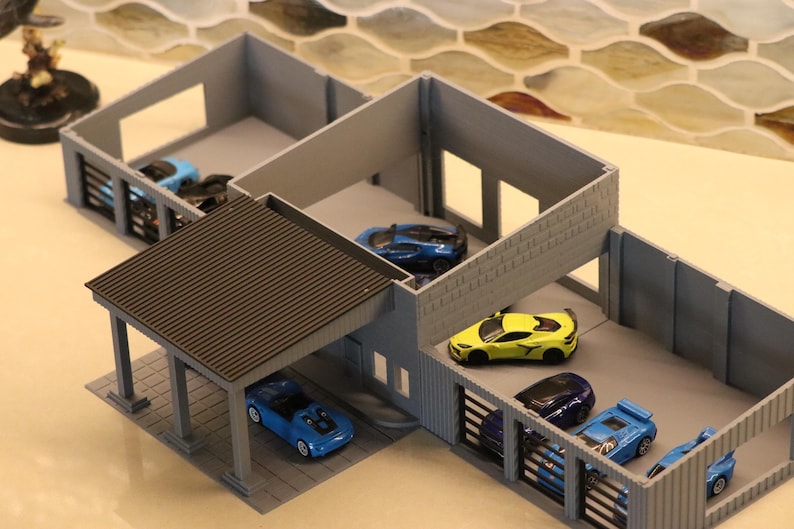 May include: A gray miniature garage structure with a brown roof, housing several toy cars in various colors, including blue and yellow. The model has open bays and a carport, designed for toy car display.
