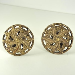 May include: Two antique brass-colored round earrings. Each earring features a central floral design surrounded by a detailed, openwork border. The earrings have a vintage aesthetic, with a slightly aged appearance.