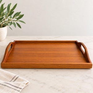 May include: A rectangular wooden serving tray with a warm brown finish and a smooth, flat surface. The tray features two integrated handles on the shorter sides, which curve upward from the base to provide a secure grip for carrying items.
