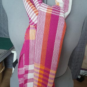 Puede incluir: Una bufanda tejida que presenta un vibrante patrón de cuadros en tonos magenta, naranja brillante y blanco. La tela muestra un tejido texturizado en forma de rejilla con largos flecos sueltos en colores a juego que cuelgan de la parte inferior.
