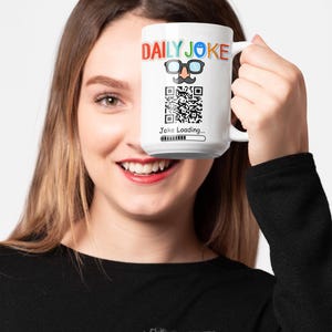 Op de afbeelding: Witte keramische mok met de tekst "DAILY JOKE" in kleurrijke letters, een cartoon snor en bril, en een QR-code. Onder de QR-code staat de tekst "Joke Loading..." met een voortgangsbalk. De mok wordt door een persoon vastgehouden.