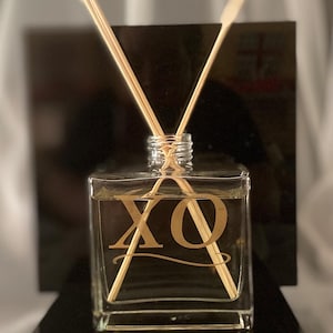 May include: A clear square glass reed diffuser bottle filled with liquid, featuring the gold letters XO printed on the front. Two light-colored wooden reeds are inserted into the neck of the bottle, extending upwards in a V-shape.