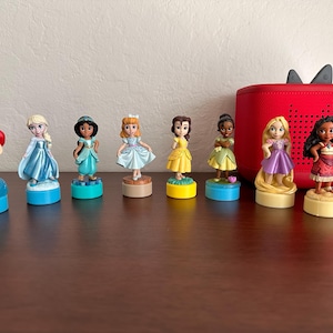 May include: A collection of eight princess figurines, each standing on a circular base. The princesses are dressed in various colorful gowns and dresses. A red speaker with black accents is in the background.