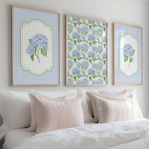 May include: Three framed art prints showcasing blue hydrangea flowers. Two prints have a blue and white striped background, the central one a floral pattern. The frames are light wood, displayed above a bed with white bedding. The room has a neutral colour scheme.