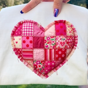 Pode incluir: Um moletom branco com um grande design de coração de patchwork. O coração é composto por vários tecidos rosa e vermelhos, incluindo bolinhas, listras e estampas florais. As bordas do coração são costuradas com uma linha decorativa visível.