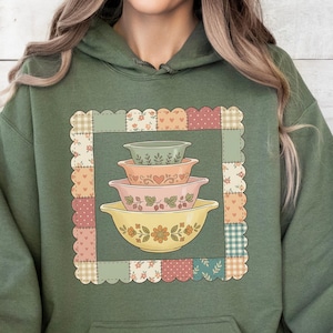 Może przedstawiać: Zielona bluza z kapturem w kolorze oliwkowym z patchworkiem i stosu mis do mieszania w stylu vintage. Miski są w odcieniach żółci, różu i zieleni, z kwiatowymi i sercowymi wzorami. Bluza ma przednią kieszeń i kaptur ze sznurkiem.