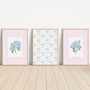 May include: Three framed art prints featuring watercolor hydrangea flowers. Two prints have a pink background with a white, scalloped border around the floral image. The third print has a repeating hydrangea pattern on a white background.
