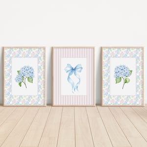 Puede incluir: Tres impresiones enmarcadas en acuarela. Dos presentan flores de hortensia azules con hojas verdes, sobre un borde con estampado floral. La tercera impresión muestra un lazo azul sobre un fondo a rayas rosas. Todo en marcos de madera clara.