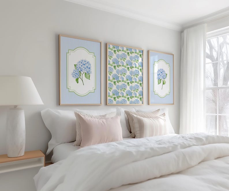 May include: Three framed art prints showcasing blue hydrangea flowers. Two prints have a blue and white striped background, the central one a floral pattern. The frames are light wood, displayed above a bed with white bedding. The room has a neutral colour scheme.