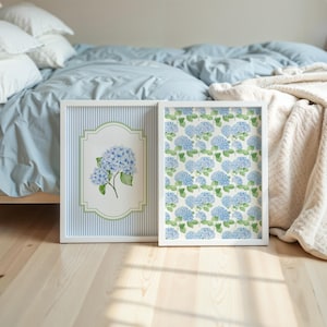 May include: Two framed art prints featuring blue hydrangea flowers. One print has a striped background with a floral illustration, the other has a repeating floral pattern. Both are in white frames.