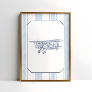 May include: A framed print featuring a blue and white biplane illustration. The artwork is set against a white background with a blue striped border and a gold frame. The biplane is detailed with propeller and wheels.