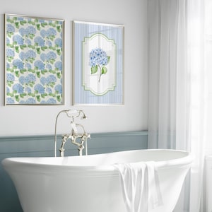 May include: Two framed floral art prints featuring blue hydrangea blooms. One print displays a pattern of hydrangeas, while the other showcases a single flower against a striped background. The prints are displayed above a white bathtub in a bathroom setting.