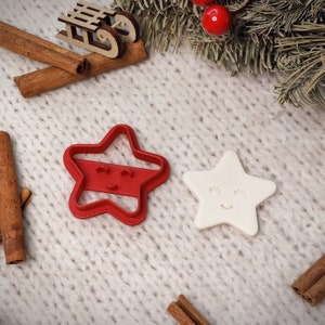 Può includere: Due stampini per biscotti a forma di stella, uno rosso e uno bianco, con facce sorridenti. Gli stampini sono su una superficie di maglia bianca.