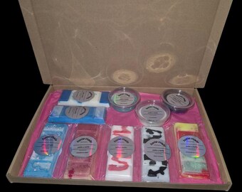 May include: A cardboard box filled with six small jars of wax melts and six larger bars of wax melts. The jars are labeled "home-made" and the bars are labeled "home-made" with different designs. The box is lined with pink paper.