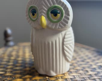 May include: A ceramic owl figurine with a cream-colored body, green and yellow eyes, and a yellow beak. The owl has a textured surface with vertical lines and is sitting on a woven surface.