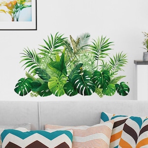 May include: A wall decal depicting a collection of tropical leaves in shades of green. The design includes large monstera leaves, palm fronds, and other foliage, creating a natural accent. The decal is positioned above a white sofa with colourful cushions.