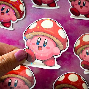 May include: A die-cut sticker featuring a pink, round character with blue eyes and a wide smile, wearing a large red mushroom cap with white spots. The sticker has a holographic finish that reflects light with small, scattered star-shaped sparkles.