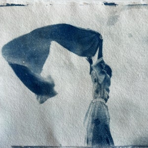 May include: A cyanotype print on textured paper featuring a monochromatic blue and white image of a figure standing with arms raised, holding a large piece of fabric that billows out to the left side, creating a sense of movement and wind.
