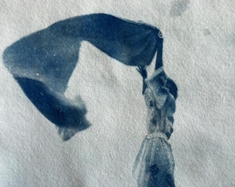 May include: A cyanotype print on textured paper featuring a monochromatic blue and white image of a figure standing with arms raised, holding a large piece of fabric that billows out to the left side, creating a sense of movement and wind.