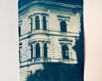 May include: A cyanotype photograph of a building with multiple windows. The building is made of white brick and has a dark blue roof. The photograph is slightly faded and has a textured surface.