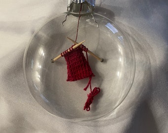Puede incluir: Un adorno de Navidad de vidrio transparente con una bufanda de punto roja en miniatura y agujas de tejer de madera en su interior.