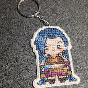 May include: A blue and brown embroidered keychain featuring a cartoon character with blue hair and a brown outfit. The keychain is attached to a silver key ring.