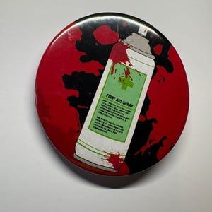 Puede incluir: Un botón redondo con un fondo rojo y negro que presenta un gráfico de un aerosol de primeros auxilios. El aerosol es blanco con detalles verdes y rojos y el texto "FIRST AID SPRAY".
