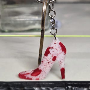 May include: A white high-heeled shoe keychain with red splatter details. The shoe charm is attached to a silver chain and key ring. The keychain is a unique accessory.