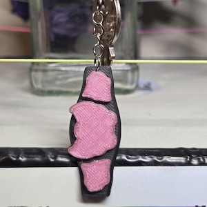 May include: A black and pink keychain featuring a championship belt design. The keychain has a silver chain and key ring. The belt has three pink sections and a black base. The design is 3D printed.