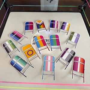Peut inclure: Collection d'autocollants en forme de petites chaises, chacun arborant un design de drapeau de la fierté différent. Les autocollants présentent une chaise pliante avec des rayures et des motifs colorés représentant diverses identités LGBTQ+. En arrière-plan, un arrangement floral et un ring de catch.