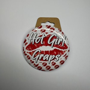 May include: A round button with a white background covered in red lipstick kiss marks. The button has the words "Hot Girl Graps" in a cursive font, with a red lipstick kiss mark in the center.
