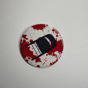 Op de afbeelding: Een ronde knoop met een witte achtergrond en een rood spatterontwerp. Een zwarte stoelafbeelding is gecentreerd op de knoop. De knoop is een pinback-stijl.