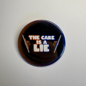 May include: A circular badge with a dark gradient background of orange, black, and blue. The text "THE CAKE IS A LIE" is in white and orange. Two small flags with cake designs are on either side.