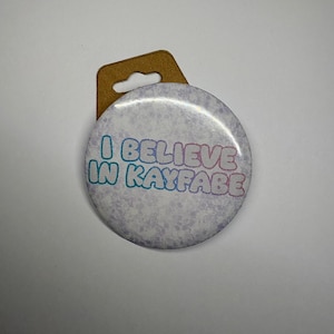 Könnte beinhalten: Ein runder Button mit dem Text "I BELIEVE IN KAYFABE" in Blau und Rosa. Der Button hat einen hellvioletten Hintergrund mit einem dezenten Blumenmuster. Er ist an einer braunen Kartonrückseite befestigt.