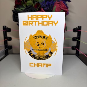 May include: A white birthday card with the words "HAPPY BIRTHDAY CHAMP" in orange. The card features a gold and black wrestling championship belt design. The card is displayed in a miniature wrestling ring.