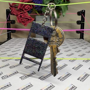 May include: A glittery black chair-shaped keychain with a silver key ring, paired with a gold-colored key. The key has the word "SEO" and "erreture" engraved on it. The keychain is a unique accessory.