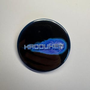 Op de afbeelding: Een ronde, zwarte knoop met het woord "HADOUKEN" in zilverkleurige, gestileerde tekst. De tekst staat tegen een blauw, wolkenachtig ontwerp. De knoop heeft een glanzende afwerking en is waarschijnlijk een verzamelobject of accessoire.