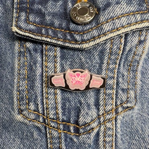 Puede incluir: Un pin de esmalte rectangular con forma de cinturón de campeonato de lucha libre con fondo negro y detalles en rosa. El centro presenta un emblema rosa estilizado con la palabra Divas escrita en fuente cursiva, sujeto a una chaqueta de mezclilla azul.