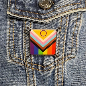 May include: A square enamel pin featuring the Progress Pride flag design, which includes horizontal stripes of red, orange, yellow, green, blue, and violet, with a chevron of black, brown, light blue, pink, and white, and a yellow circle with a purple border.