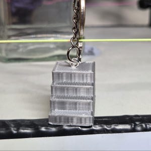 Può includere: Un piccolo portachiavi grigio stampato in 3D con un design a scala in miniatura. Il portachiavi è attaccato a un anello e una catena di metallo argentato. La scala ha quattro gradini visibili. Lo sfondo è sfocato, con una linea gialla e un contenitore di vetro.