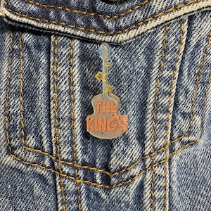 May include: A guitar-shaped acrylic pin featuring the orange text THE KING'S centered on the body. The design includes small yellow star accents along the neck and headstock, with a translucent finish that reveals the blue denim fabric underneath.