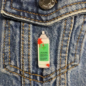 Puede incluir: Un pin de esmalte rectangular con forma de aerosol, con etiqueta verde claro, cuerpo blanco y salpicaduras de pintura roja. La etiqueta muestra el texto FIRST AID SPRAY y texto instructivo. El pin está sujeto a una tela de mezclilla azul.